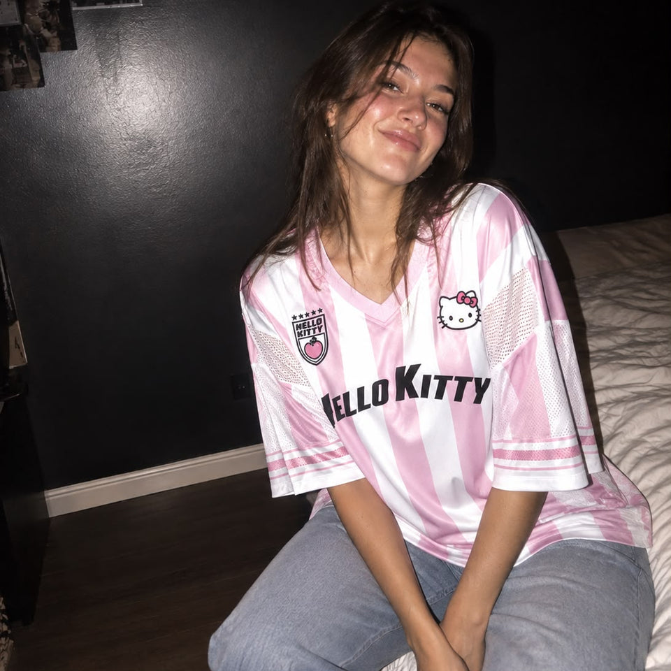 A woman sitting on a bed wearing a pink and white striped Hello Kitty soccer jersey and light blue jeans in a dimly lit bedroom.